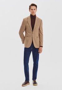 Brązowa i beżowa marynarka w wzór houndstooth na ciemnym golfie, zestawiona z niebieskimi dżinsami i brązowymi butami, stojąca na tle białej ściany.