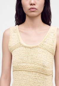 Femme portant un haut tricoté sans manches de couleur crème avec des motifs texturés, montrant ses épaules et le haut de sa poitrine sur un fond uni.