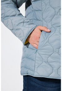 Chaqueta acolchada de color azul claro con una superficie texturizada, que cuenta con un bolsillo lateral y un forro amarillo de contraste visible en el puño.