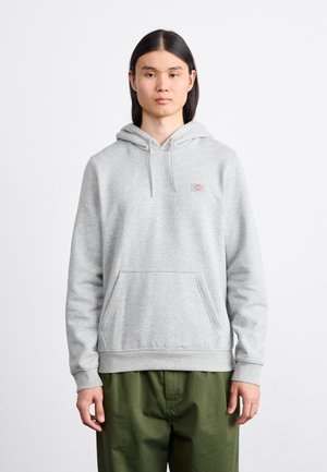 Un sweat à capuche gris en mélange de coton, avec une poche ventrale, des poignets côtelés, et un petit logo. Associé à un pantalon vert à coupe ample.