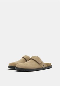 Beige velúr slip-on cipők, emelt hátsó résszel és széles pánttal a tetején. Fekete gumitalp, texturált tapadással.