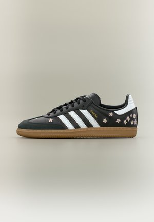 Baskets Adidas Samba noires avec des bandes blanches, une semelle marron, de petits motifs floraux roses et blancs, et le texte "SAMBA" en doré sur le côté.