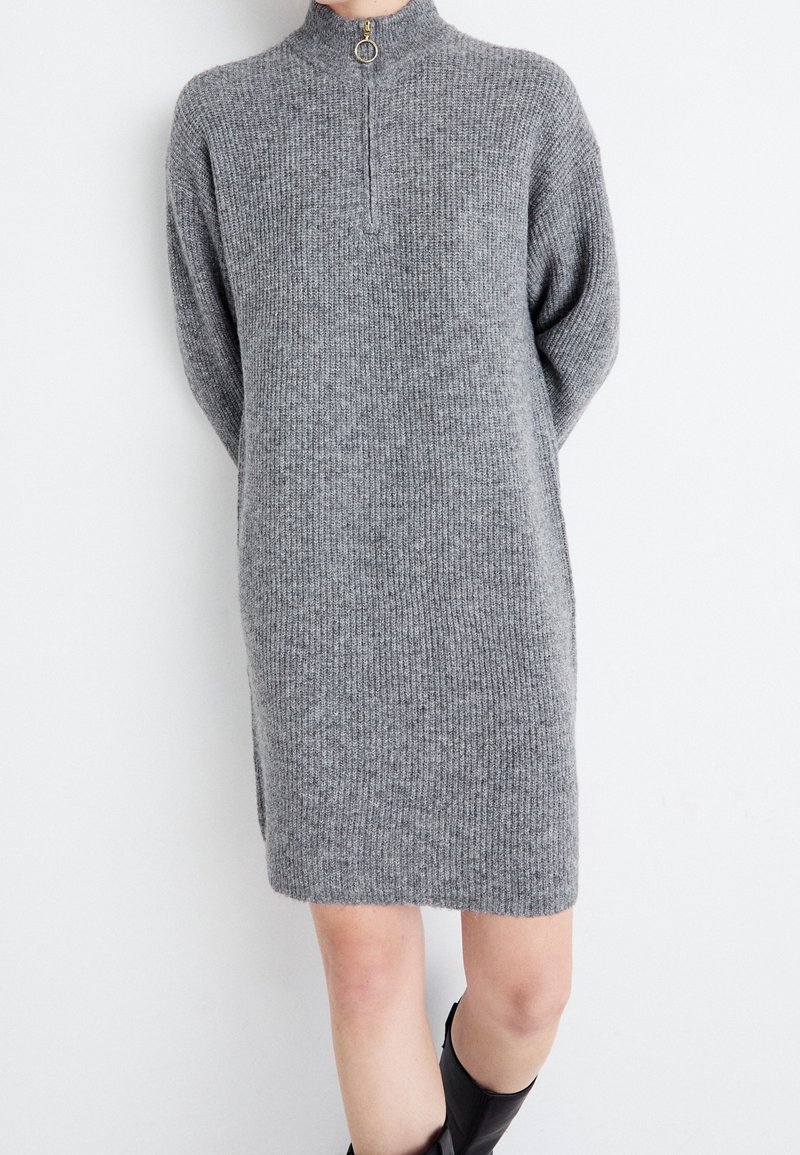Robe en maille côtelée grise longueur genou avec manches longues et col à fermeture éclair devant, portée avec des bottes noires.
