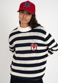 Persona joven con gorra roja y suéter a rayas azul marino y blanco con un logo rojo en el pecho, de pie frente a un fondo liso.