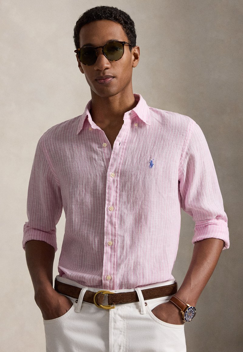 Mees kannab roosade triipudega nööpidega särki, valgeid pükse, pruuni vööd, käekella ja tumedaid päikeseprille, seisab käed taskutes.