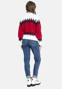 Kobieta ubrana w sweter z czerwonym, białym i czarnym wzorem, niebieskie jeansy z podwiniętymi nogawkami oraz białe sznurowane botki, stojąca tyłem.