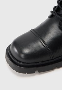 Sort læderstøvle med syet cap toe, tyk gummisål med groft mønster og sorte snørebånd på en lys baggrund.