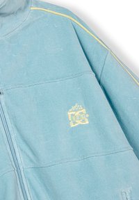 Giacca a zip di colore azzurro chiaro realizzata in tessuto morbido, con piping giallo come dettaglio e un piccolo logo ricamato sul lato sinistro.