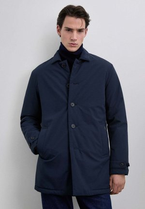 Joven con un abrigo azul marino abotonado sobre un suéter de cuello alto oscuro, de pie contra un fondo gris claro y liso.