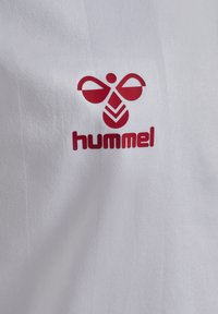 Weißes Sporthemd mit einem roten Hummel-Logo, das ein abstraktes Design und Text aufweist, aus strukturiertem Material gefertigt.