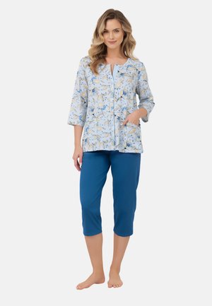 Kobieta stojąca boso, ubrana w jasnoniebieską kwiecistą bluzkę zapinaną na guziki z rękawami do połowy przedramienia oraz jednolite, niebieskie spodnie do połowy łydki.