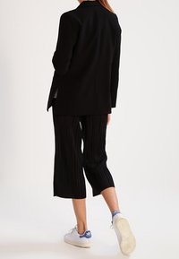 Blazer noir avec une coupe ajustée, associé à un pantalon cropped plissé noir. Baskets blanches avec des accents bleus visibles aux talons. Texture lisse.