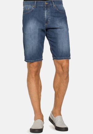 Uomo che indossa pantaloncini di jeans blu di media lunghezza e scarpe slip-on grigie, in piedi davanti a uno sfondo semplice.