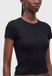 Woman with dark skin and braided hair wearing a fitted black short-sleeve ribbed shirt, shown from shoulders to waist against a plain background.
