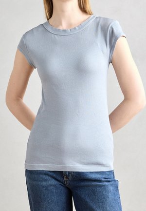 Vrouw draagt een lichtblauw aansluitend T-shirt met korte mouwen en een blauwe spijkerbroek, staand met handen achter haar rug tegen een effen achtergrond.