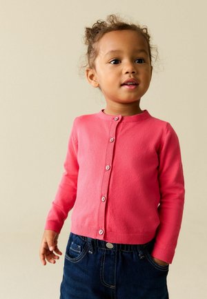 Kleinkind trägt eine rosa Strickjacke mit Knöpfen und dunkle Jeanshose, steht mit einer Hand in der Tasche und schaut leicht zur Seite.