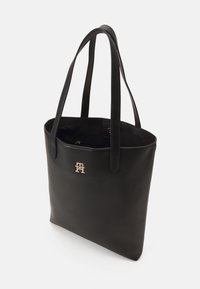 Sac en cuir noir avec deux longues poignées, arborant un accent logo doré à l'avant et un design intérieur spacieux.