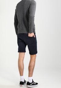 Grijze textuurtrui, marineblauwe opgerolde shorts, witte sokken, zwarte sneakers. Uitzicht van achteren dat de pasvorm en kleuren van de kleding benadrukt.