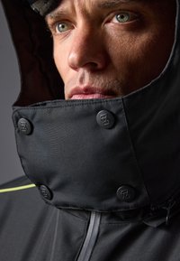 Chaqueta de tela negra con un alto cuello y botones. Presenta un material texturizado, costuras visibles y un detalle de cremallera en la parte delantera.
