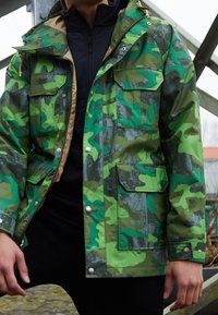 Green camouflage waterproof jacket with multiple pockets, a hood, and a zip closure. Features a contrasting tan interior and smooth texture.