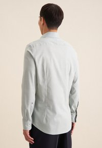 Maglietta a maniche lunghe grigio chiaro con colletto, polsini abbottonati e una texture liscia. Presenta una linea dritta e una piega sul retro.