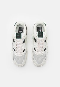 Zapatillas blancas y grises con materiales de malla y cuero, con acentos en negro, cordones planos y etiquetas de logo en la lengüeta y el costado.