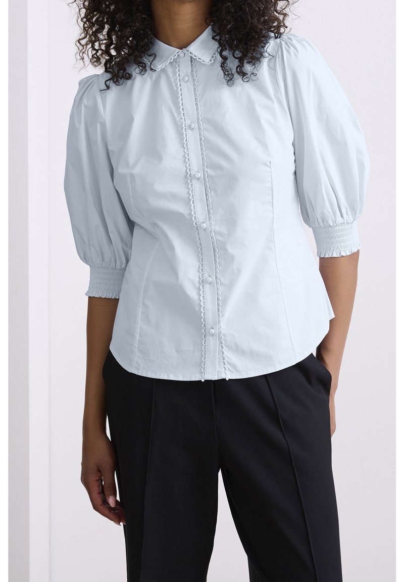 Light blue puff-sleeve blouse with a collared neckline, front button closure, decorative trim, and smocked cuffs. Paired with black pants.