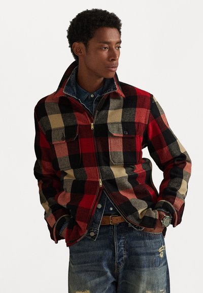 Red, black, and beige plaid flannel jacket with two chest pockets, closed zipper, and collar. Worn over a denim shirt and distressed jeans.