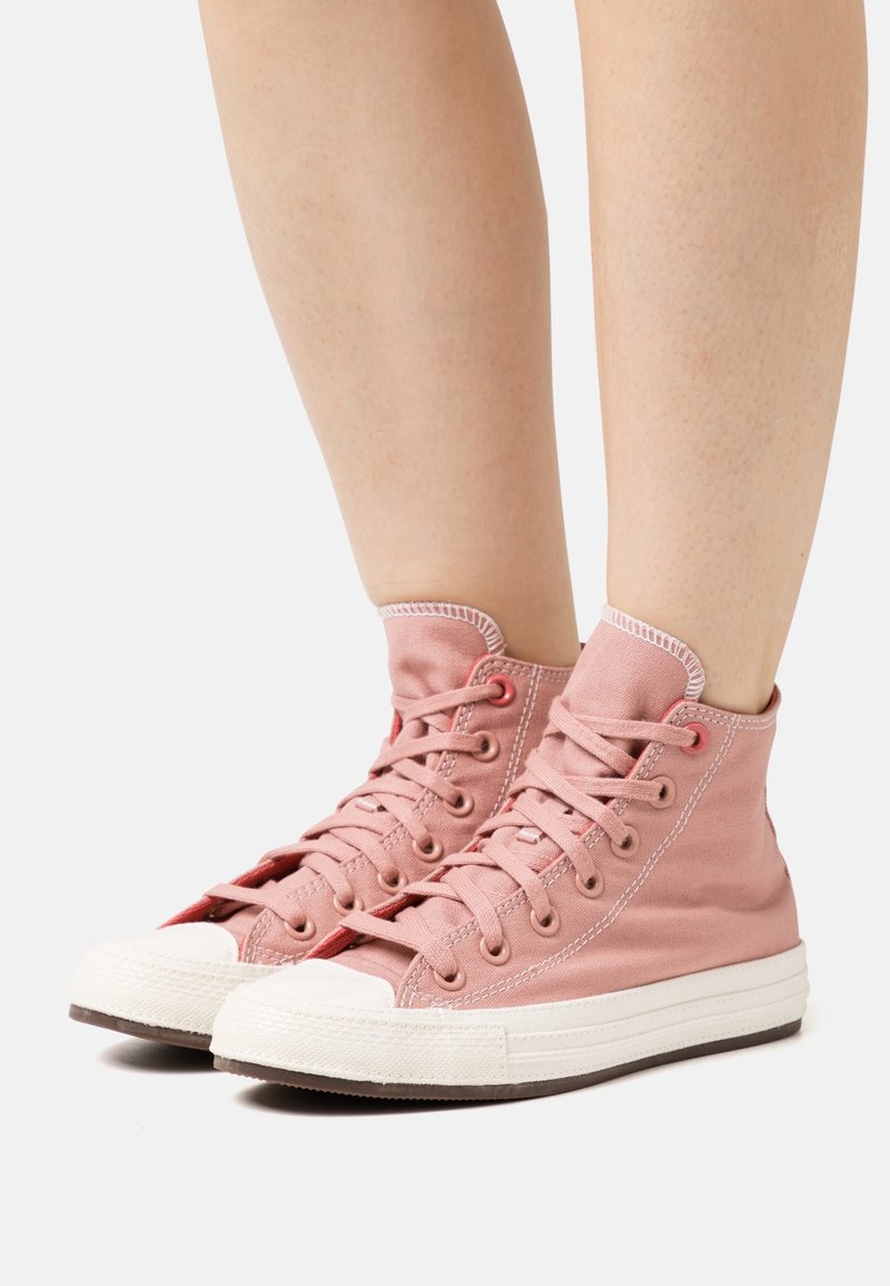 Jambes portant des baskets montantes en toile rose avec semelles et lacets en caoutchouc blanc, présentées sur un fond clair uni.