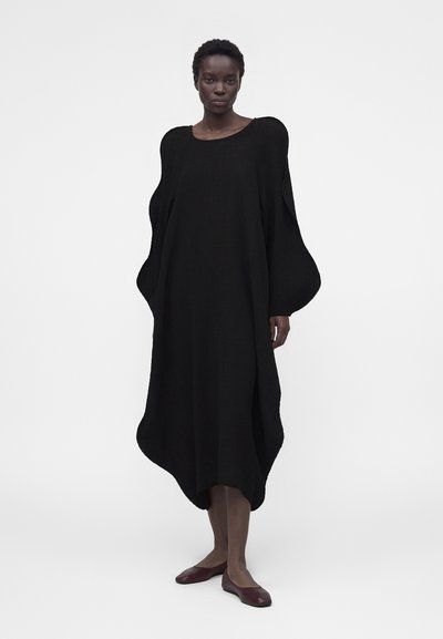 Γυναίκα που φοράει ένα φαρδύ, μαύρο φόρεμα με μακριά μανίκια, με υπερμεγέθη φουσκωτά μανίκια και στρογγυλεμένη μυσή, σε συνδυασμό με καφέ επίπεδα παπούτσια.