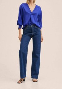 Królestwo niebieska bluzka z dekoltem w serek i luźnymi rękawami, w połączeniu z wysokim stanem, prostymi ciemnymi dżinsami i czarnymi sandałami na obcasie z odkrytymi palcami.