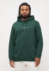 Bărbat cu dreadlocks, purtând un hanorac verde închis cu buzunar frontal și pantaloni albi, stând pe un fundal simplu și deschis la culoare.