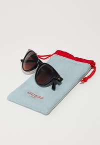 Occhiali da sole neri con lenti marroni sfumate, caratterizzati da un design elegante e piccoli accenti decorativi, poggiati su un sacchetto blu con cordino rosso.
