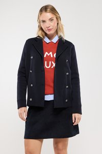 Veste marine à double boutonnage avec des boutons en argent, superposée sur un sweat-shirt rouge avec du texte blanc, et une chemise à col bleu clair.