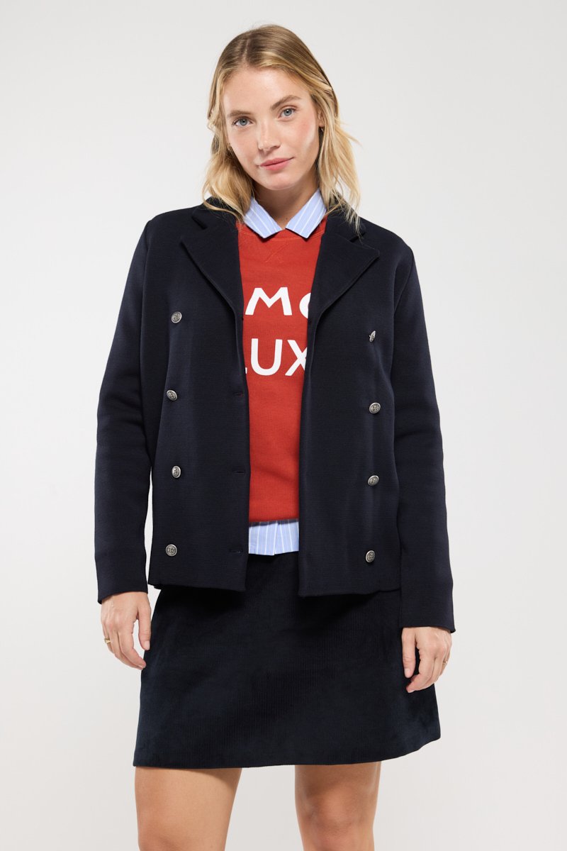 Veste marine à double boutonnage avec des boutons en argent, superposée sur un sweat-shirt rouge avec du texte blanc, et une chemise à col bleu clair.