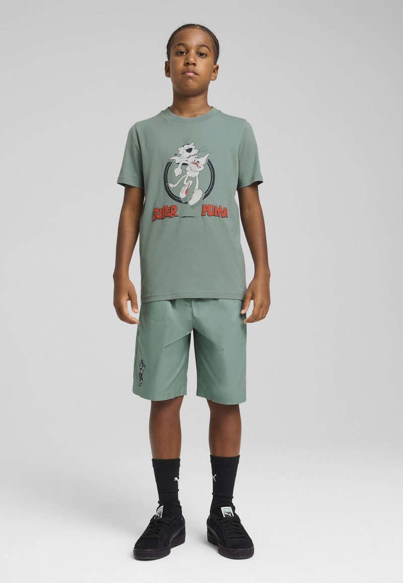 T-shirt en coton vert clair avec un motif graphique et du texte rouge, associé à un short vert clair, porté avec des chaussettes et des chaussures noires.