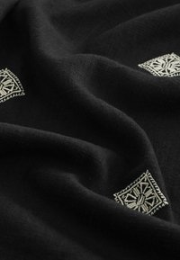 Tissu noir avec des plis doux présentant de petits motifs géométriques en losange blancs brodés, espacés sur toute la surface.