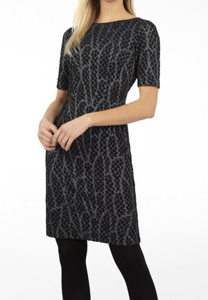 Femme portant une robe noire et grise à manches courtes, longueur genou, avec des collants noirs et un bracelet en argent.