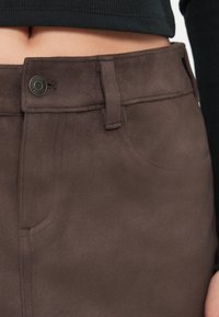 Fustă din suede sintetic maro, cu o textură netedă, buzunare frontale și detaliu cu buton metalic la talie, având o siluetă ajustată.