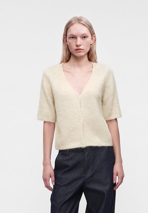 Jeune femme portant un cardigan crème à manches courtes et duveteux avec un col en V profond et un pantalon sombre taille haute sur un fond gris uni.