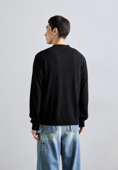Schwarzer Strickpullover mit Rundhalsausschnitt, gerippten Bündchen und Saum. Getragen mit hellblauem Denim-Hose mit aufgesetzten Taschen.