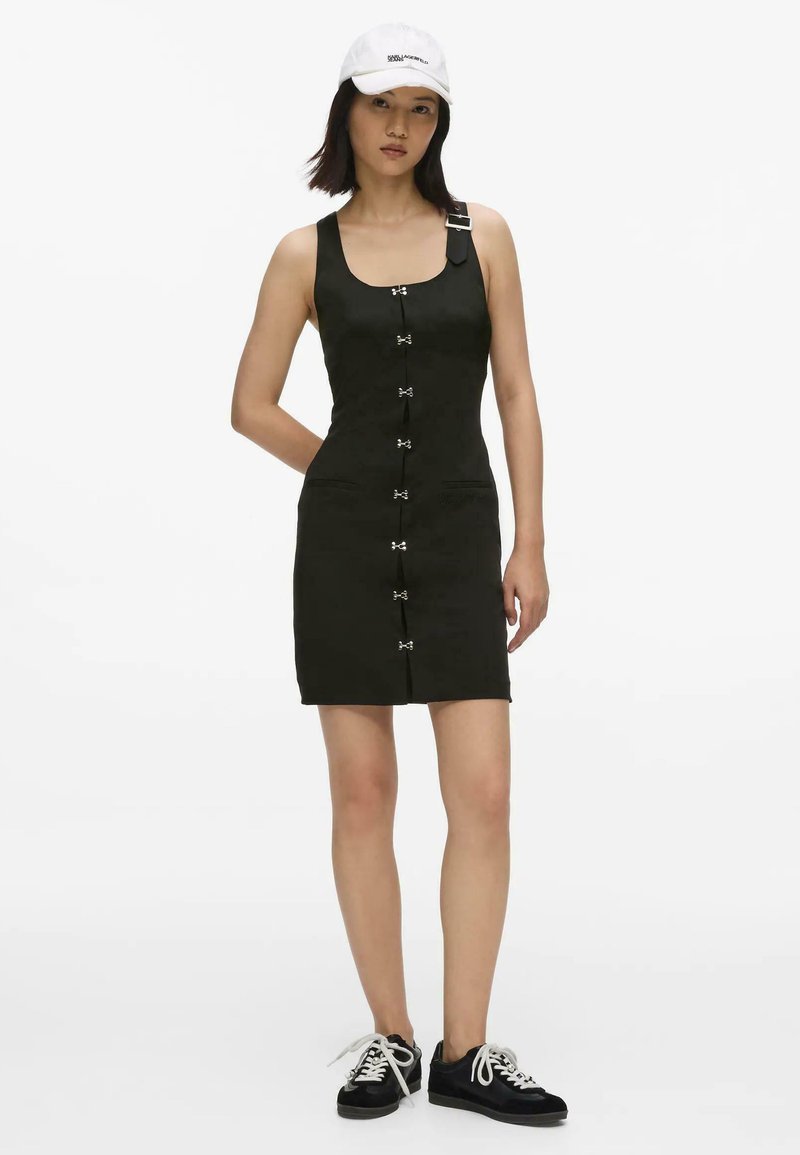 Femme portant une mini-robe noire sans manches avec des fermoirs devant, une casquette blanche et des baskets noires avec des lacets blancs, debout avec une main dans le dos.