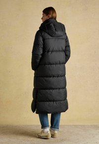 Dlouhý černý péřový kabát s kapucí, s prošívaným designem a bočními knoflíky. Nosený přes džíny a světle zbarvené tenisky.