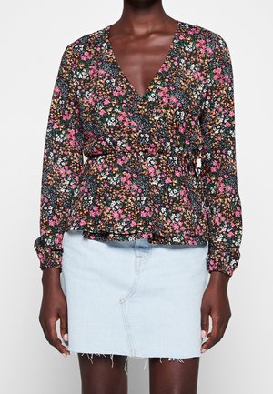 Kvinde iført en sort, blomsterpræget langærmet slå-om-bluse og en lyseblå denim mininederdel med frynset kant, stående mod en hvid baggrund.