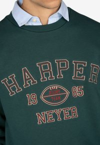 Felpa verde con colletto a coste; presenta il testo "HARPER RUGBY TEAM 1985" e un design con una grafica di un pallone da rugby in accenti rossi e beige.