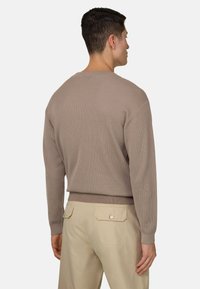 Homme de dos portant un pull à manches longues côtelé taupe et un pantalon beige avec des poches arrière boutonnées, debout devant un fond clair.