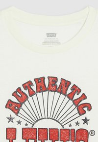 Camiseta de algodón blanca con un gran gráfico rojo de 'AUTÉNTICO' con un diseño de estallido solar y contornos negros. La etiqueta muestra la marca y los detalles de fabricación.