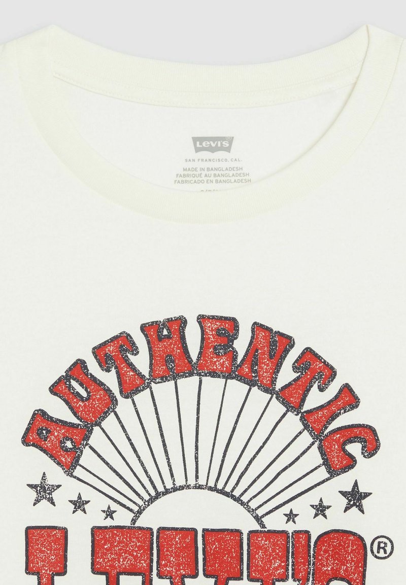 T-shirt en coton blanc avec un grand graphisme rouge 'AUTHENTIC' au design en rayons de soleil et contours noirs. L'étiquette indique les détails de la marque et de la fabrication.