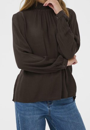 Woman wearing a dark brown long-sleeve blouse with gathered neckline and cuffs, paired with blue denim jeans, posing with crossed arms.