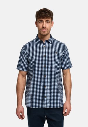 Mand iført en kortærmet, blå og hvid ternet skjorte med knapper, stående mod en ensfarvet lys baggrund.